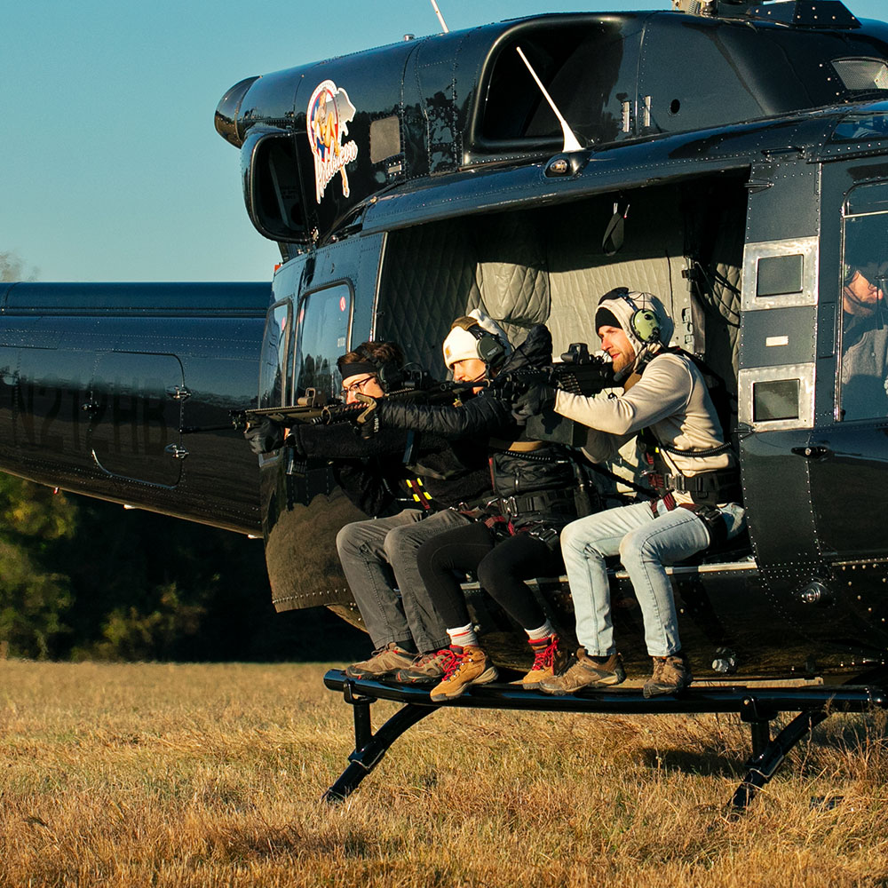 Three people sitting on the edge of a helicopter on the ground shooting machine guns