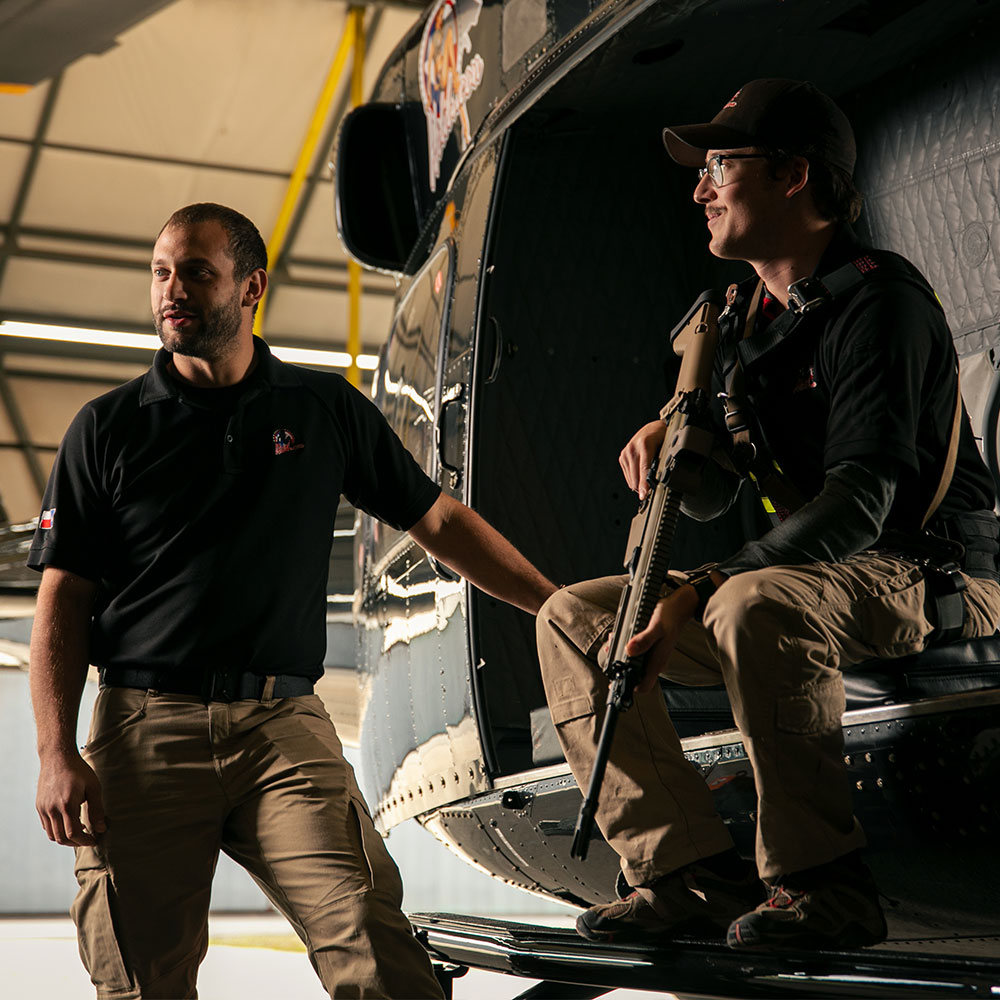Man teaching safety for Helibacon experience next to a man holding a machine gun to demonstrate